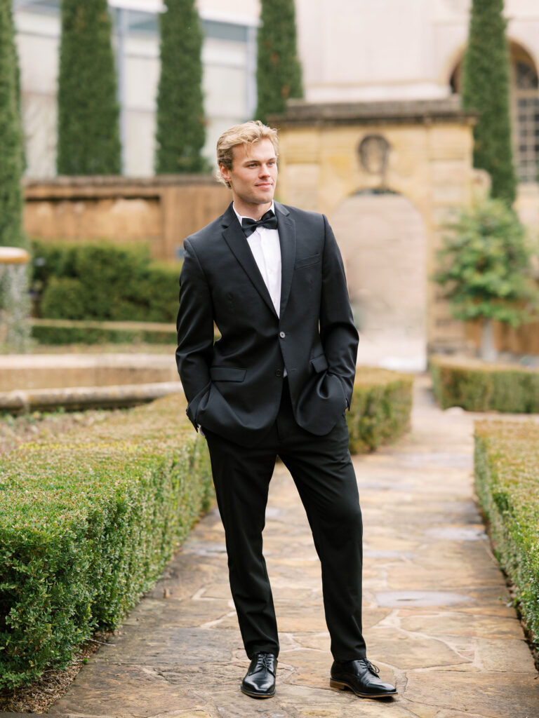 Groom stands in European style gardens in classic black tuxedo.