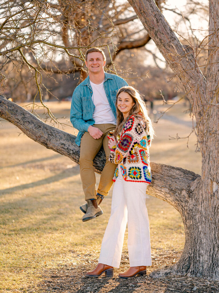 A couple takes a photo in front of a tree at City Park in Denver. The man is sitting in a branch in the tree, and his wife is standing in front of him, leaning on his leg.