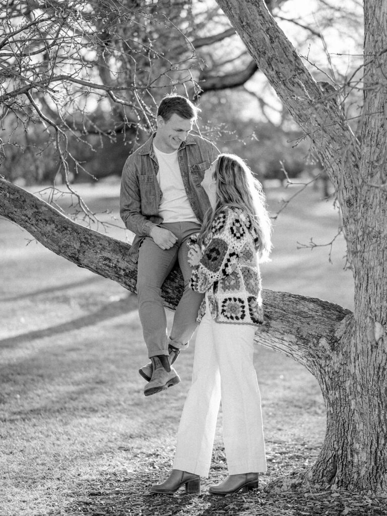 A couple takes a photo in front of a tree at City Park in Denver. The man is sitting in a branch in the tree, and his wife is standing in front of him, leaning on his leg.