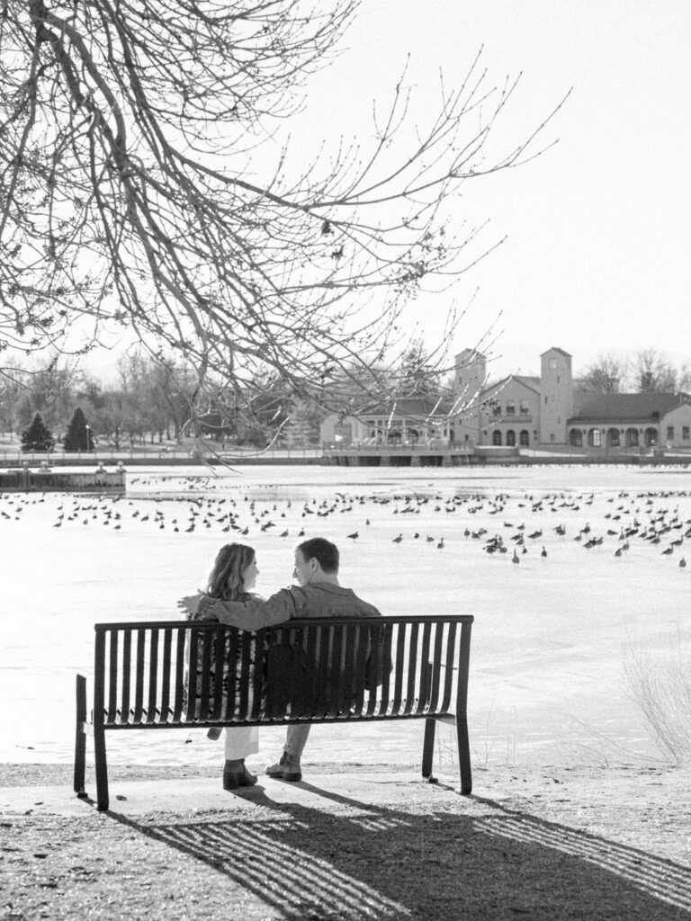 A couple sits on a bench in front of a pond at City Park in front of the Downtown Denver skyline.
