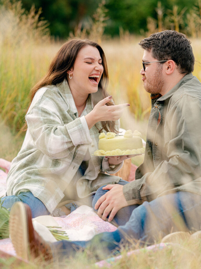 A couple sits on a picnic blanket in a park. The girl is holding a cake, laughing. She has just put a fingerful of icing on her fiance's nose.