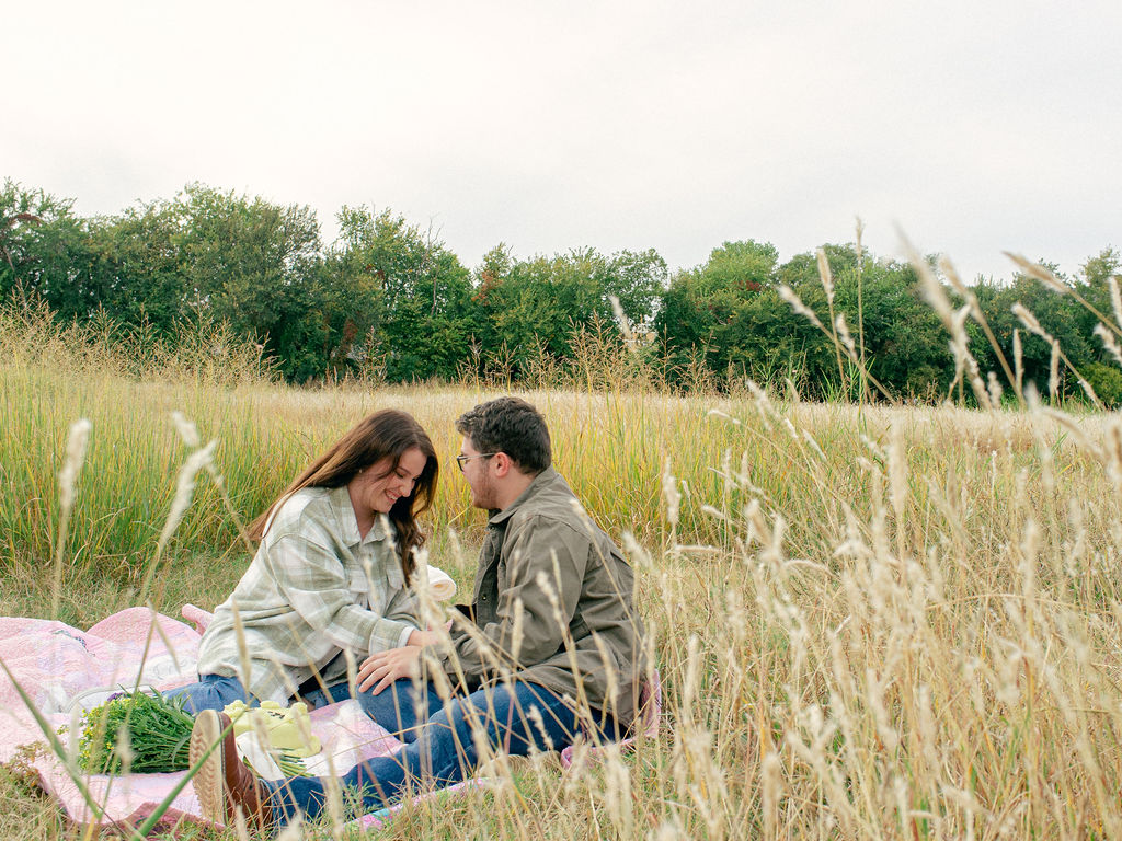 A couple sits on a picnic blanket. They are looking at each other and they have a cake in front of them.