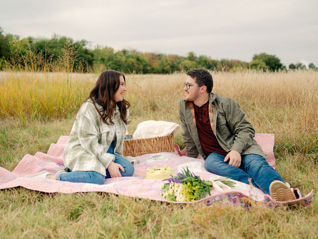 A couple sit on a picnic blanket, looking at each other.