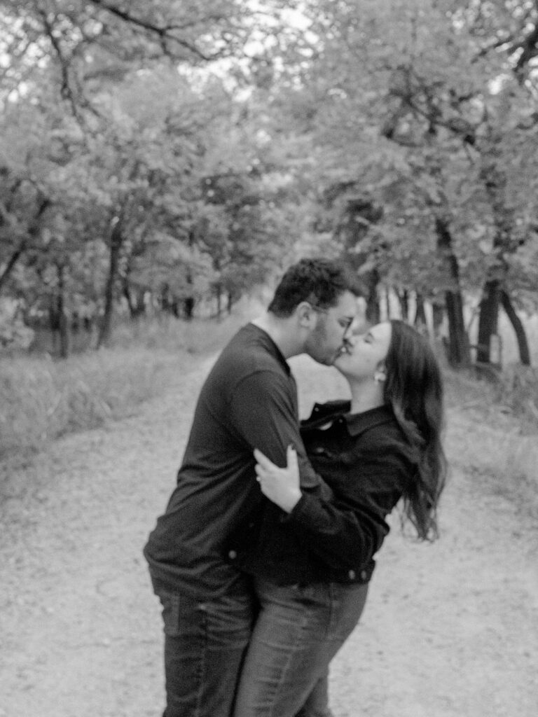 A couple kiss on a gravel path surrounded by autumn trees.