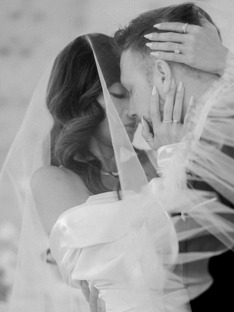 A bride and groom embrace nose to nose under a ruffled tulle veil.