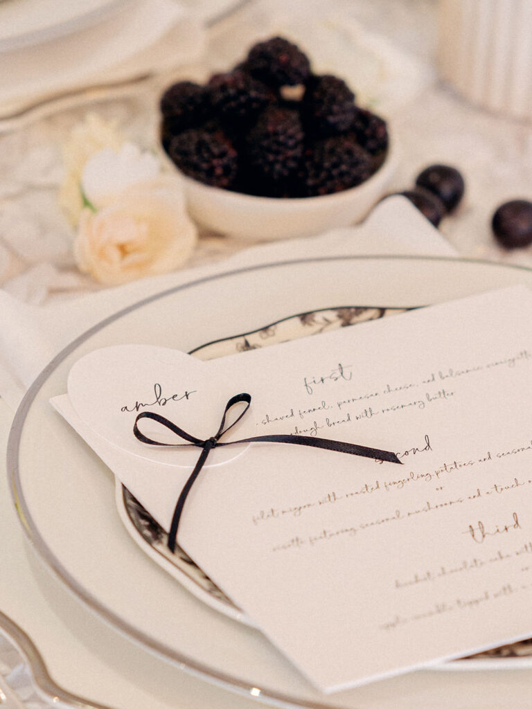 A close up of a place setting on a wedding reception tablescape with a bespoke menu featuring a black bow on the placecard.