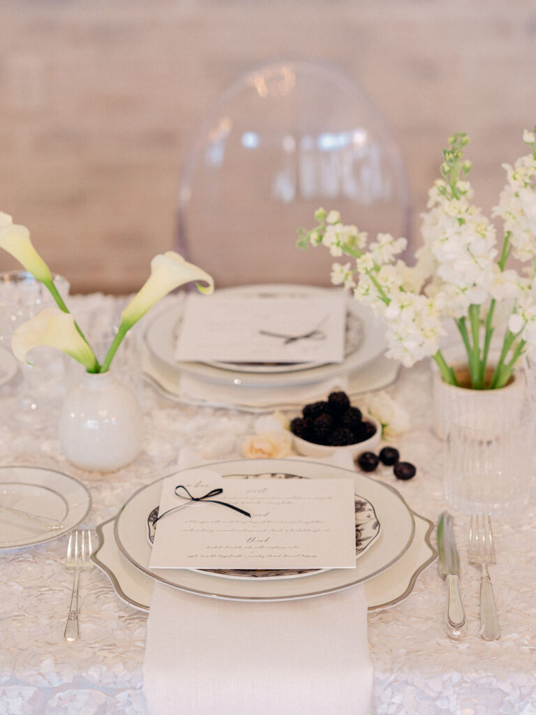 A wedding tablescape with black and white plates, a menu with a bow on it, fruit and white flower centerpieces.