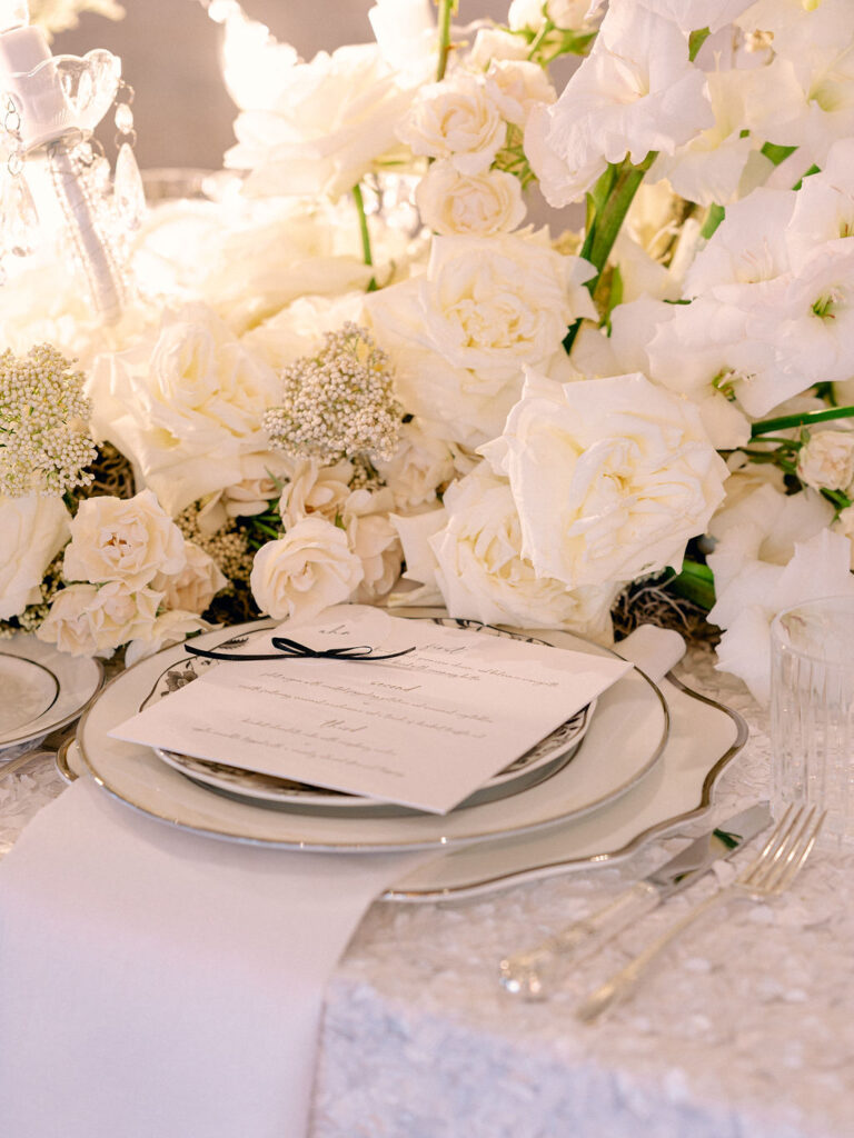 A place setting on a table at a wedding reception. Surrounded by white flowers and candle light, a menu with a bow on it sits on top of a black and white plate setting.