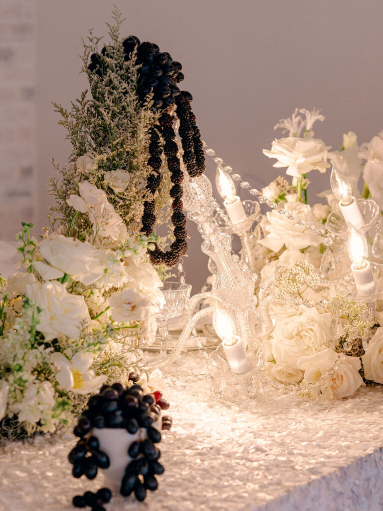 An elaborate black and white centerpiece on a wedding tablescape with loads of white flowers and blackberries surrounding a lit chandelier sitting on the table.