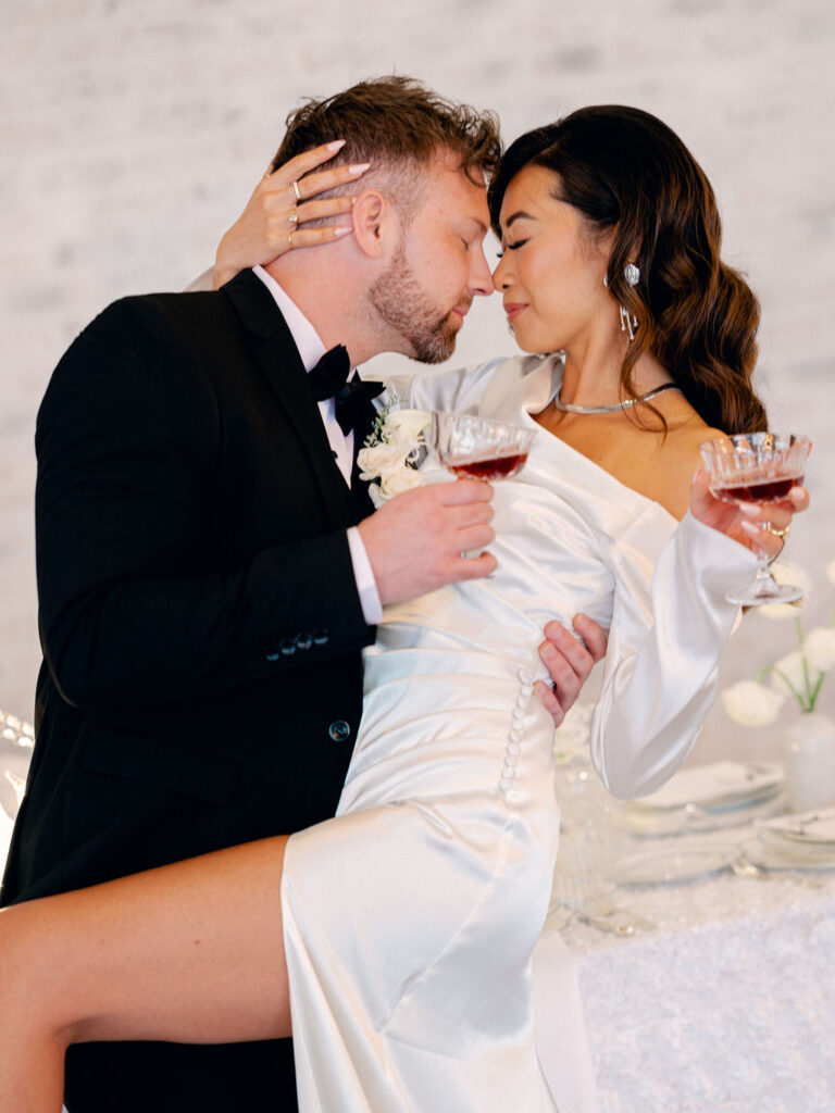 A couple embraces each other, nose to nose. They are holding red wine in their glasses.