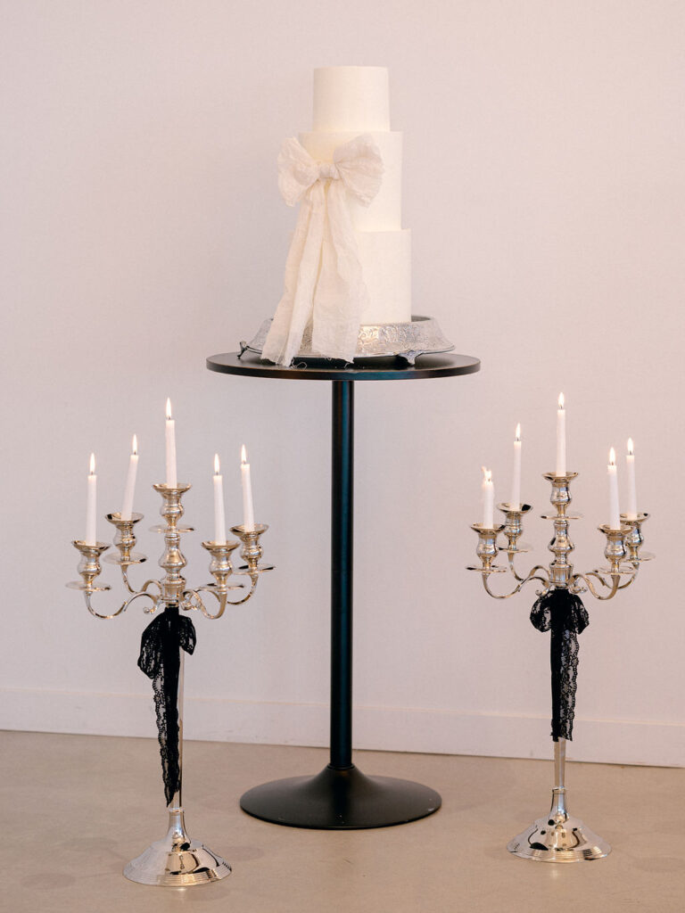 A white wedding cake with a large bow on the middle tier stands on a table between two large candelabras with black bows tied around them.