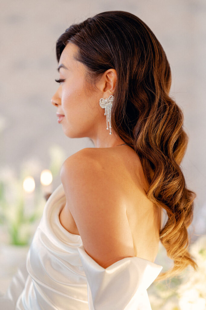 A bride stands with her back to the camera. She is wearing a modern satin gown, with curls in her hair, and a statement bow earring.