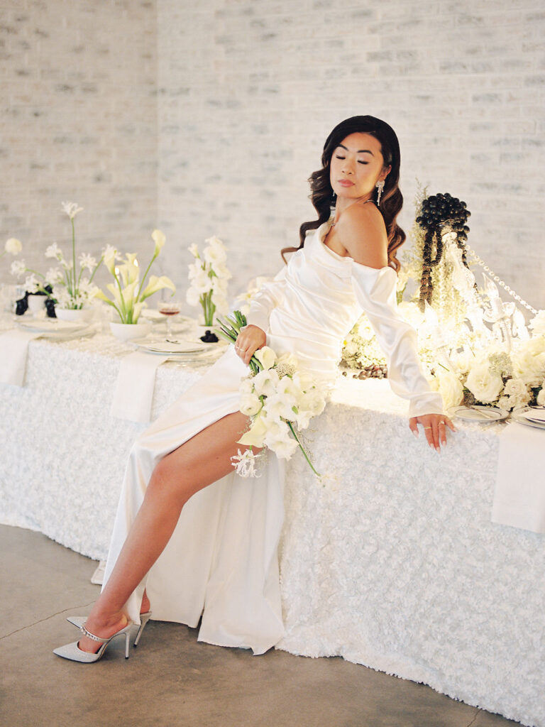 A bride in a modern satin wedding gown with a slit in the leg leans against a table at the wedding reception decked in black and white florals and fruit.