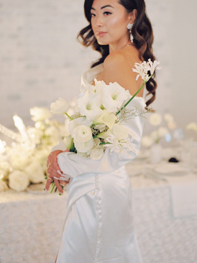 A bride in a modern satin gown holds a bouquet of elegant white flowers in the crook of her arm.