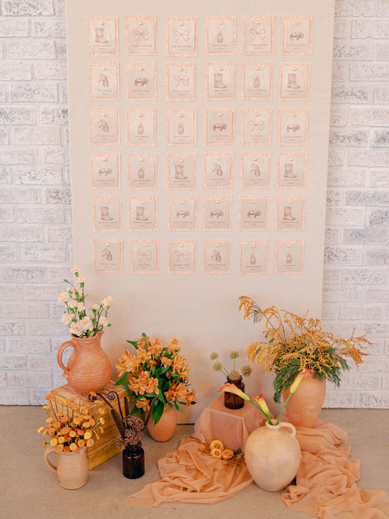 A peach toned escort card wall with orange florals in front of it.