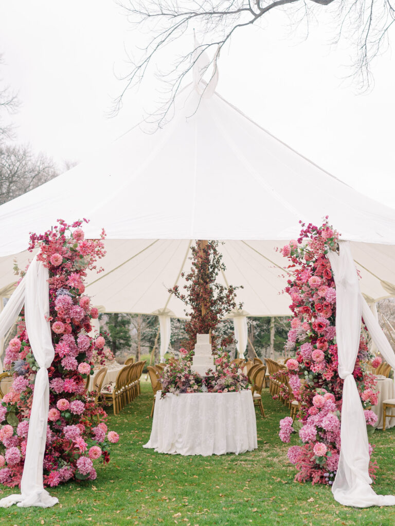 A tented wedding reception with a white, textured wedding cake surrounded by a pink floral installation.