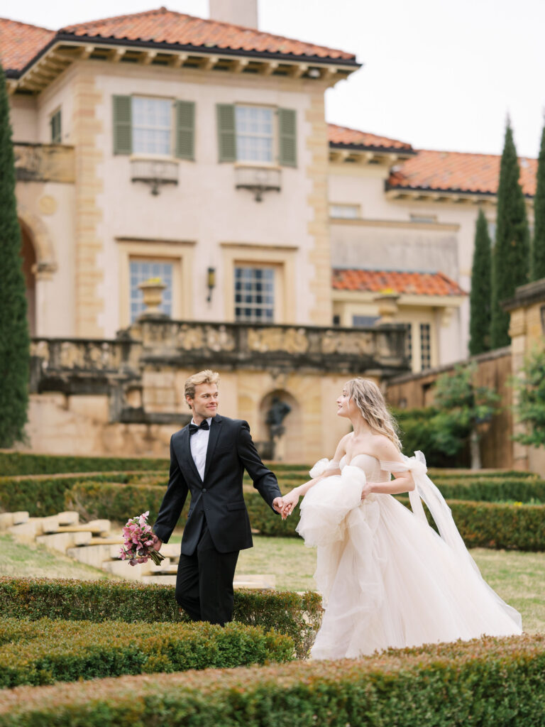 Groom leads bride by the hand as they walk through European style gardens at a historic musuem.