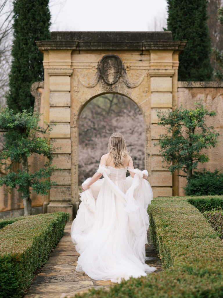Bride runs through arch way in European gardens with dress flowing behind her.