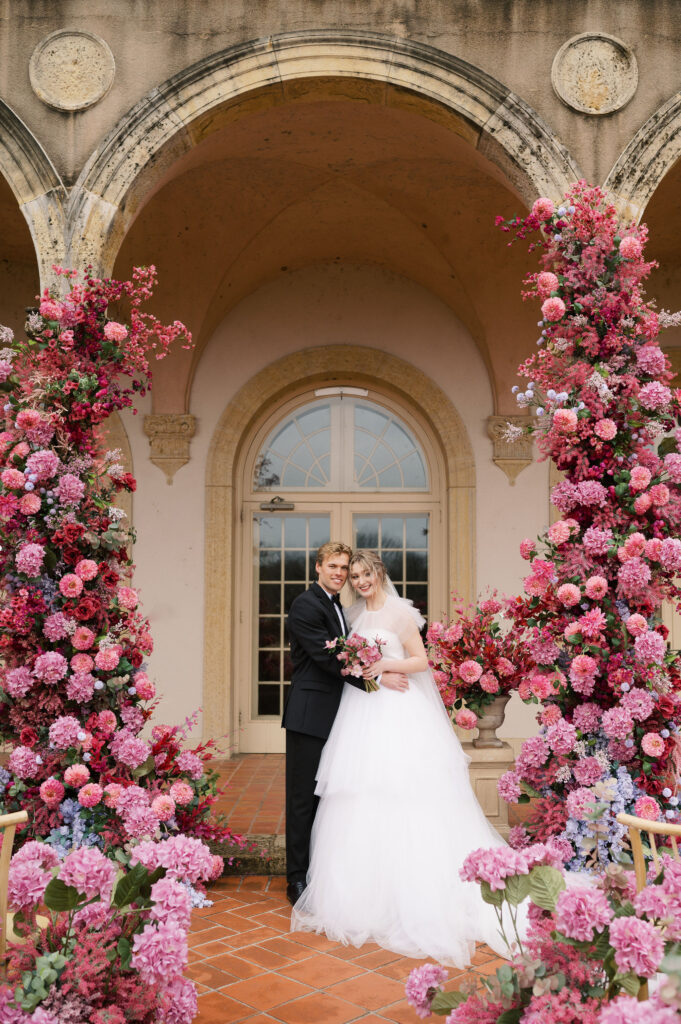 Bride and groom smile under intricate pink floral ceremony arch.