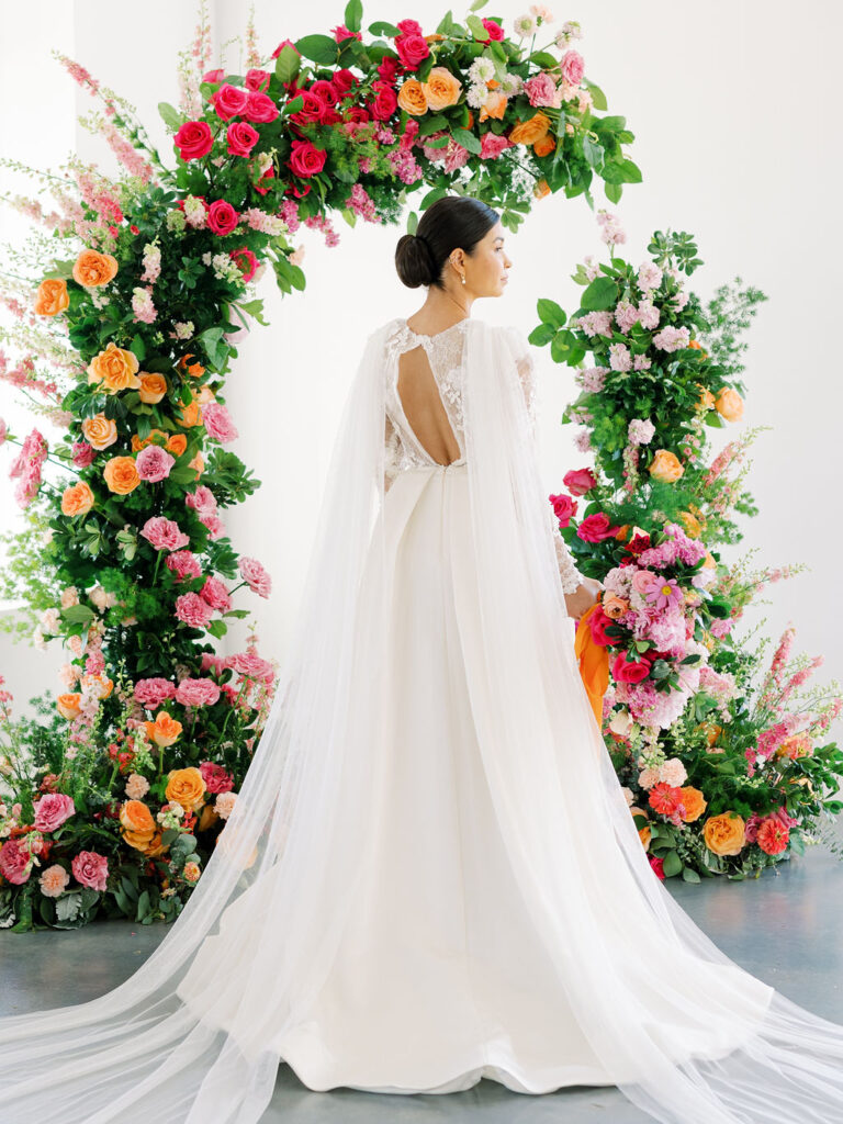 Bride stands with her back turned to the camera under pink and orange floral arch. She is wearing a dress with a cape and looking off to the right.