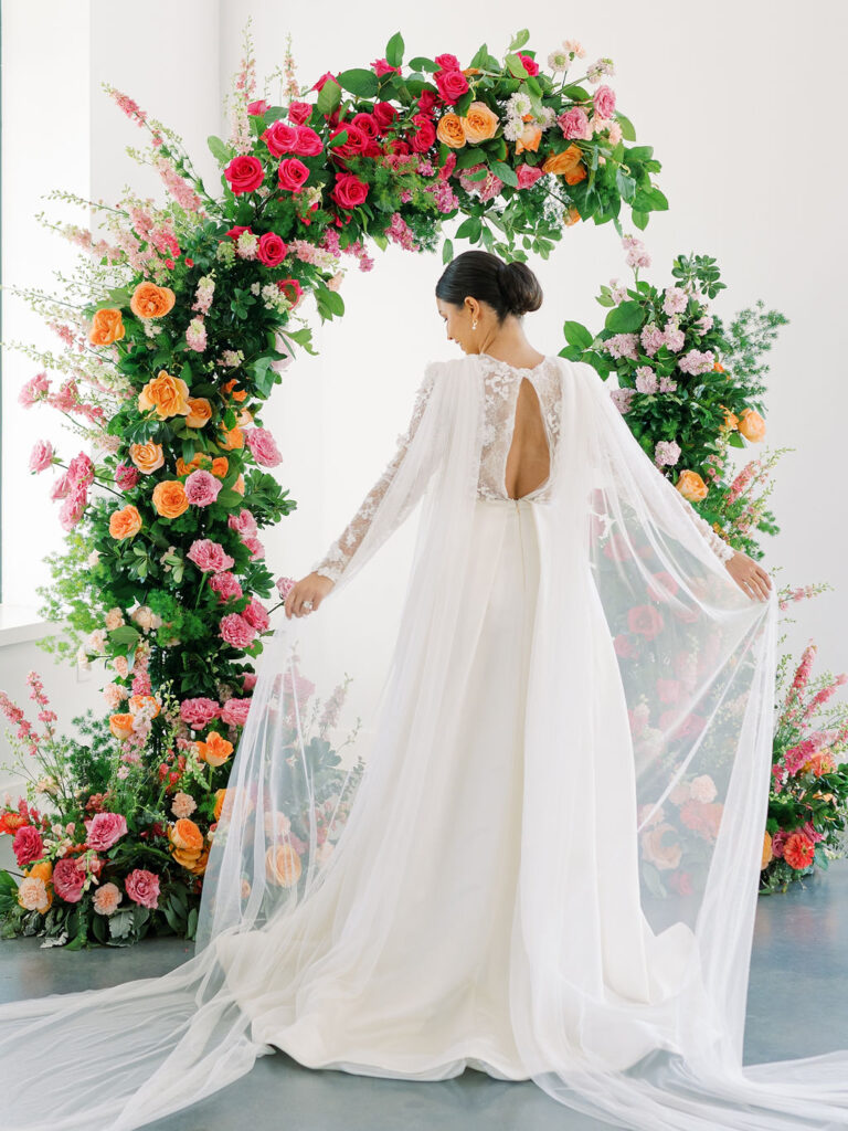 Bride standing under pink and orange floral arch has her back turned to the camera. She holds the cape of her dress with either hand and is twirling slowly.