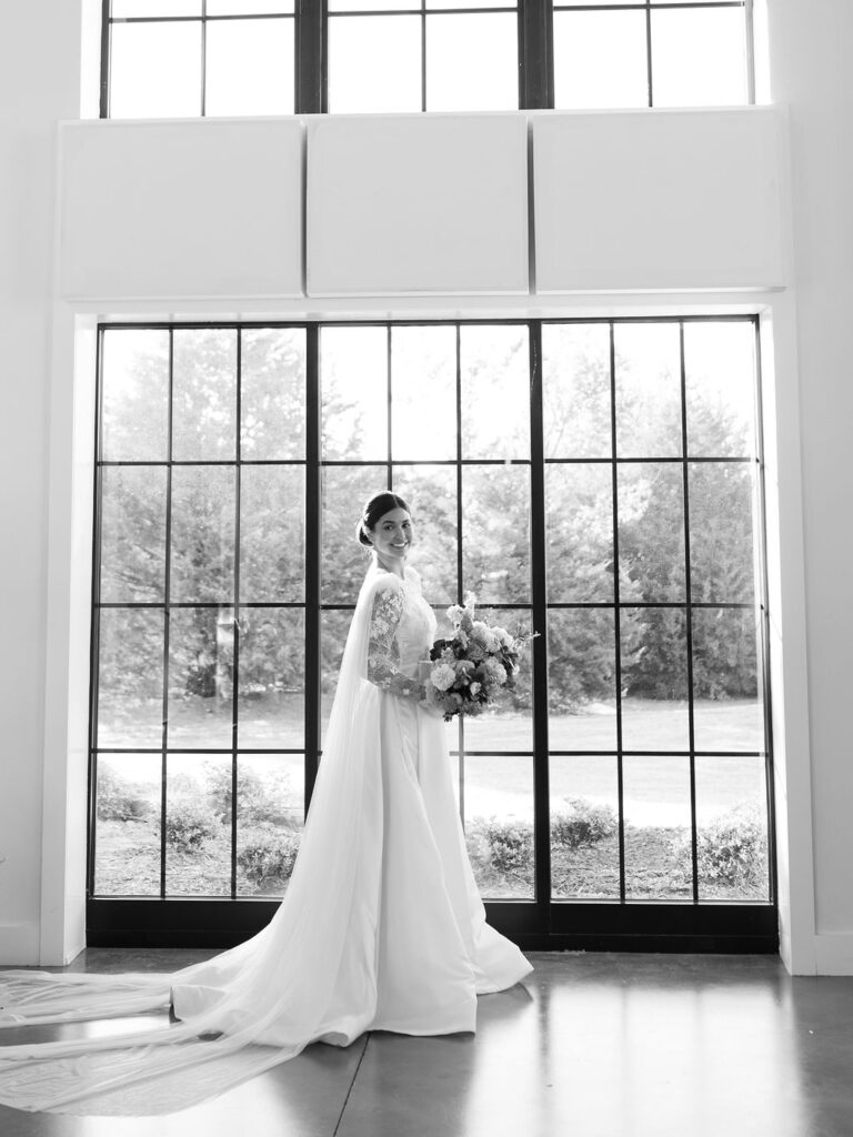 A bride stands in front of a window in a couture wedding gown. She is holding a bouquet and looking at the camera.