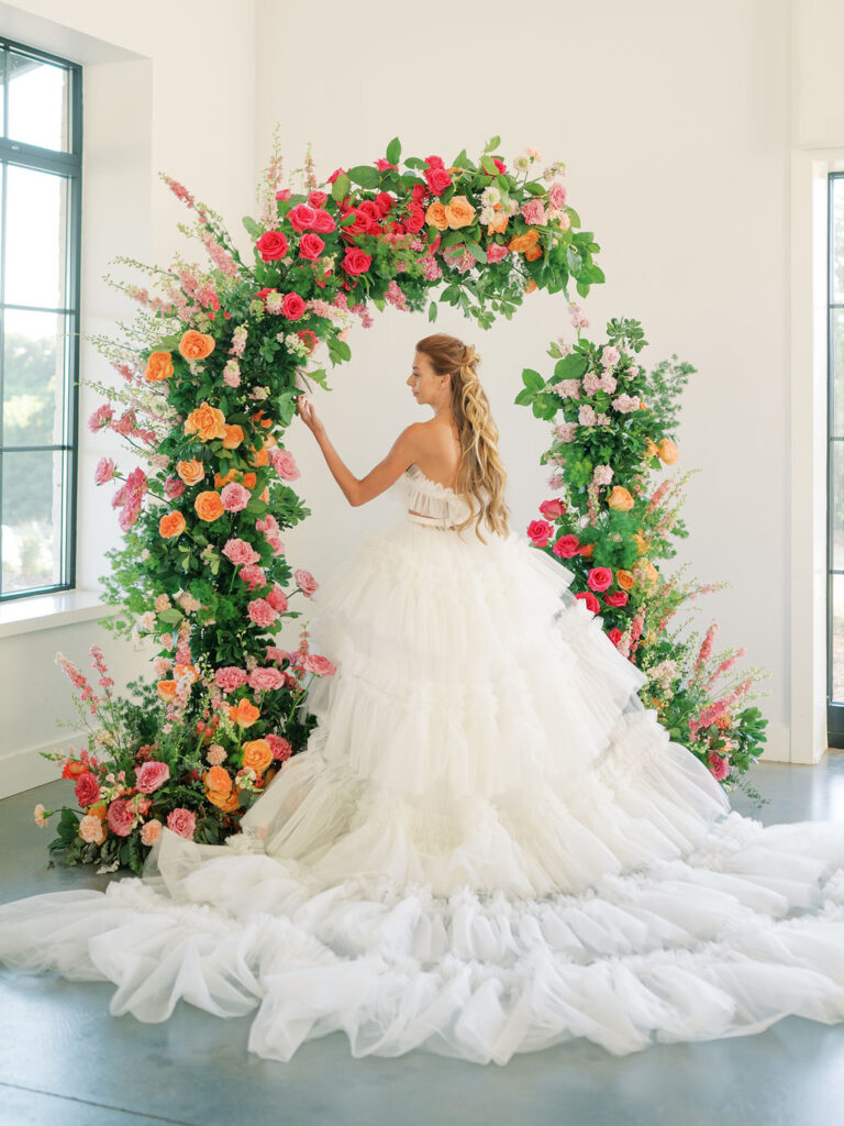 A bride with her back turned to the camera stands under a pink and orange floral arch in a ruffled dress with a long train.