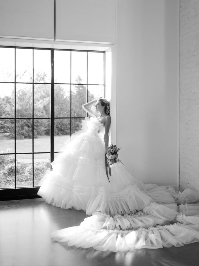 A black and white image of a bride standing in front of a window. She is leaning against a wall with her left hand bent over her head, and a bouquet in her right hand.