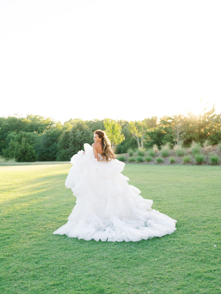 A bride in a couture dress with a long ruffled train runs away from the camera in a green field.