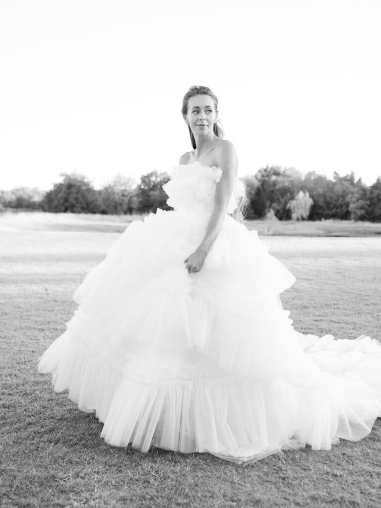 A black and white image of a bride in a custom couture wedding gown with a ruffled skirt. She is holding her skirt in one hand and looking off over her shoulder.