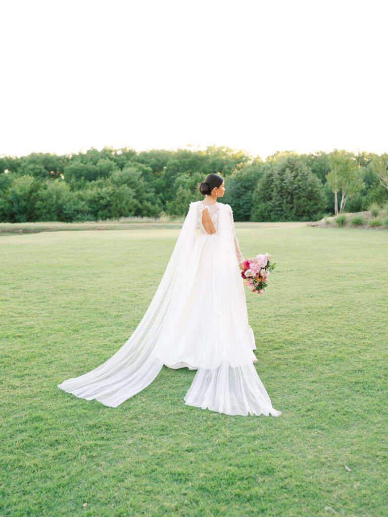 A bride wearing a custom dress with a cape that drapes over either shoulder walks away from the camera in a green field.