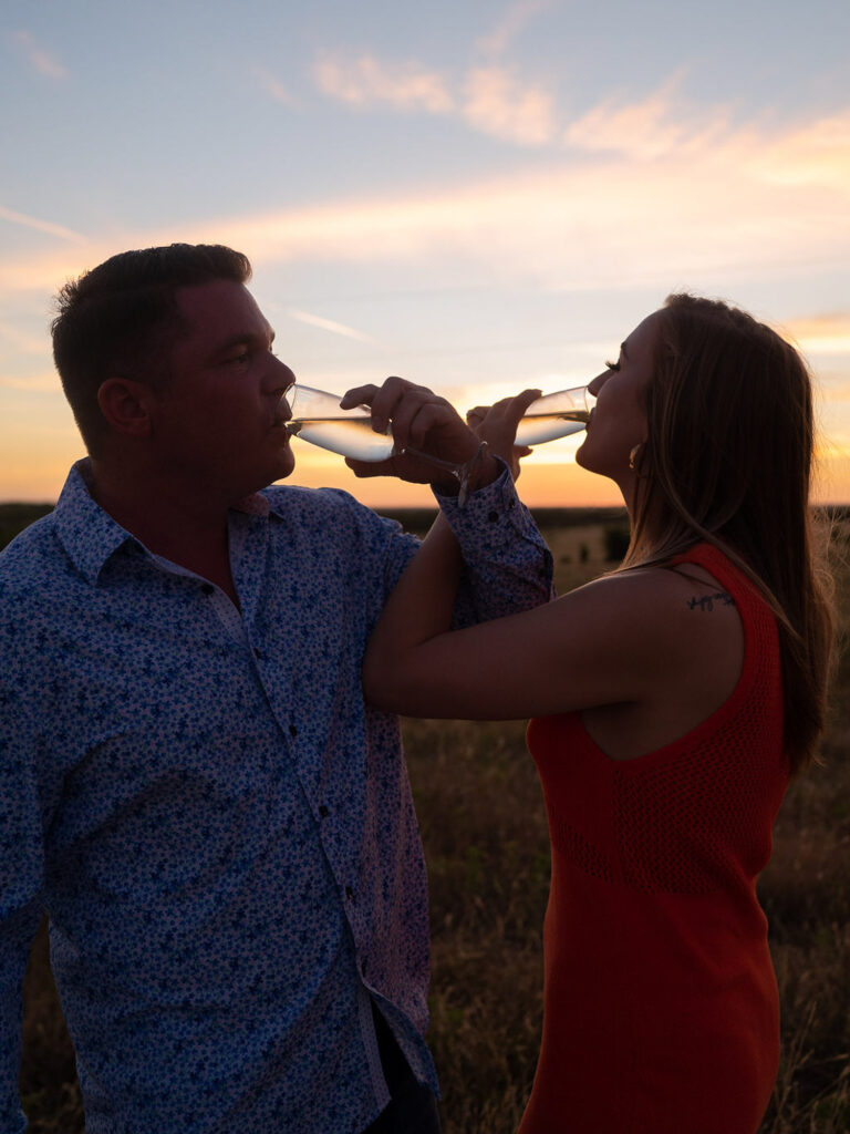 Couple links arms, drinking champagne in front of sunset on family ranch.