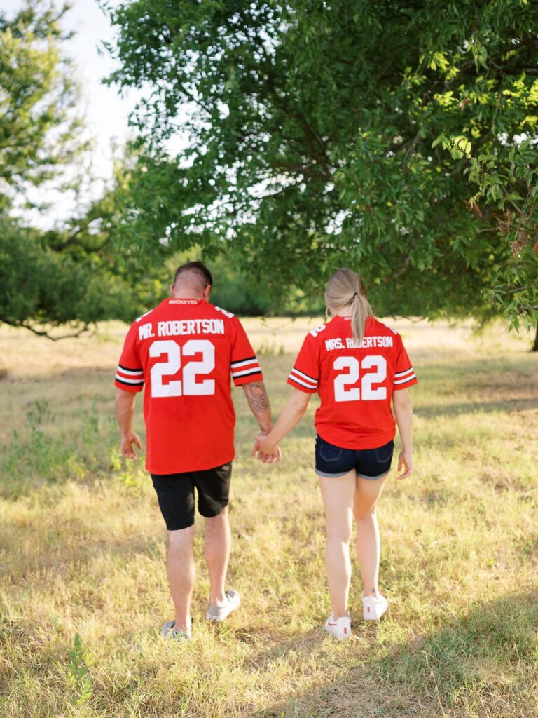 Couple holds hands, walking away from camera in open field.