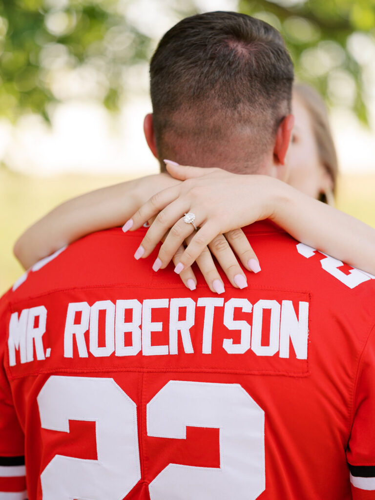 Bride wraps her arms around grooms neck with engagement ring showing, the back of his jersey reads "Mr. Robertson"