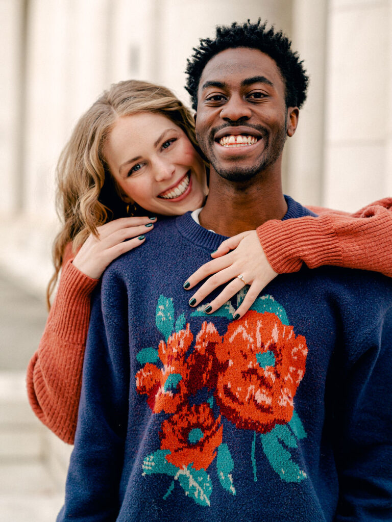 A couple stands in front of a white pavilion in a park. The guy, in a blue sweater with red flowers printed on it, smiles at the camera, while his fiance hugs behind him, her head peeking over his shoulder.