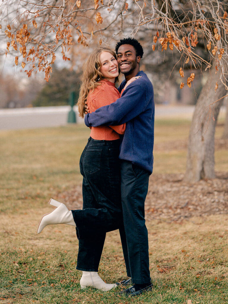 A couple hugs each other as they stand under a tree in the park. They are both smiling at the camera, and the girl has her leg kicked up behind her.
