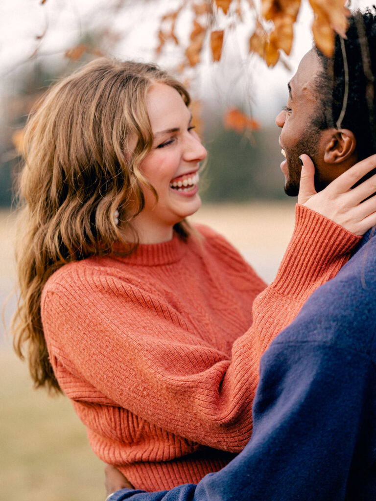 A couple embraces each other under a tree in a park. The guy is turned away from the camera, but the girl is facing it, and laughs with her hand holding his jaw.