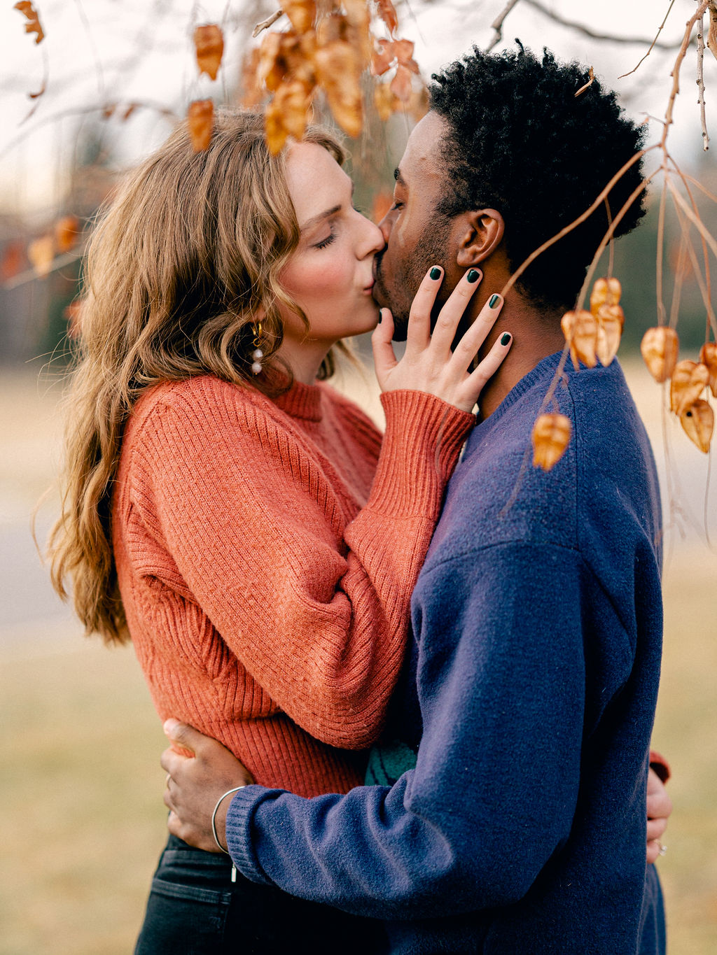 A couple kisses, holding each other close under a tree in a park. It is winter, so the leaves on the tree are brown.