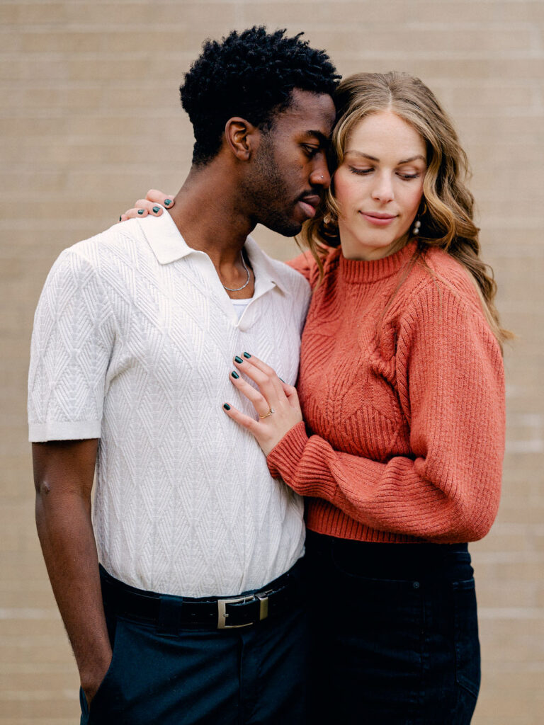 A couple stands in front of a white wall. The guy on the left, is facing the camera with his hand in his pocket, but his face is turned to the right, nuzzled against his fiance. She is hugged close to him, and is looking down her shoulder.
