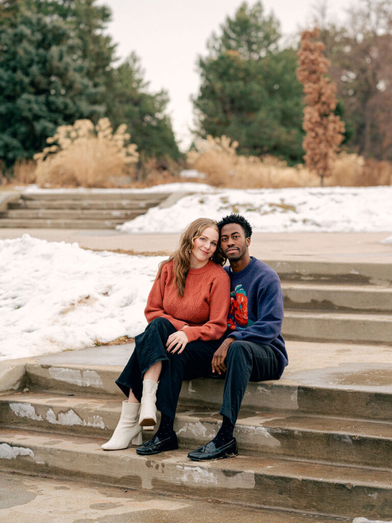 A couple sits on stairs in a park. Their arms are around each other and there's snow on the grass around them.