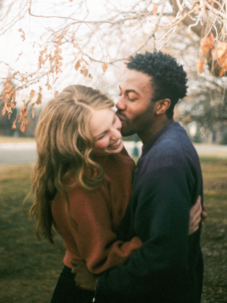 A couple embraces under a tree with browning leaves. He is kissing her cheek, and she is laughing and leaning away from him.