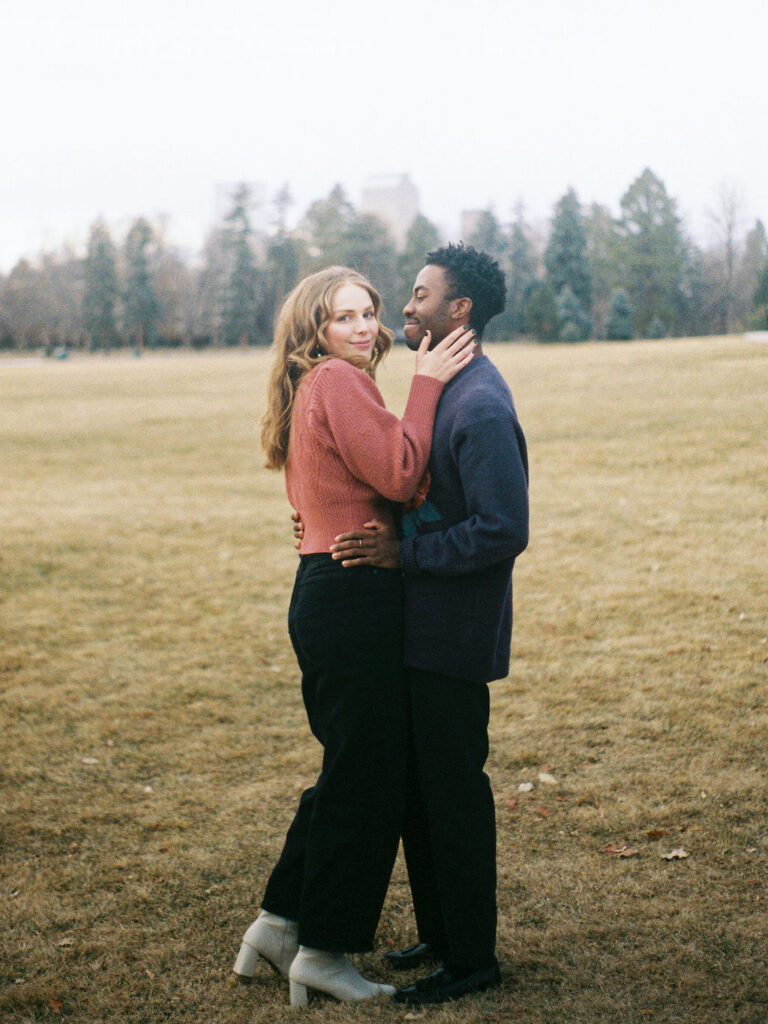 A couple stands in an open field of a park. Evergreen trees are in the distance behind them. They are embracing each other, and he is looking at her, while she smiles softly at the camera.