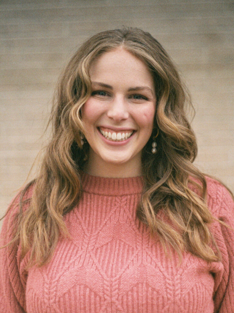 A woman stands in front of a white brick wall. It is a closeup photo of her face, and she is smiling at the camera.