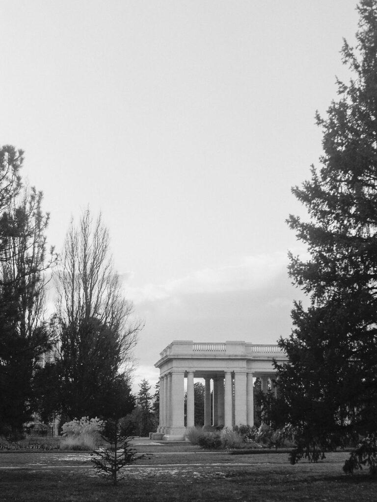 A black and white photograph of a marble pavilion in a park, surrounded by evergreen trees.