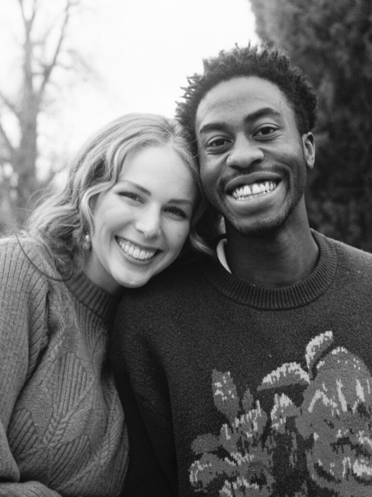 A couple smiles at the camera in a black and white photograph with their cheeks pressed close together.