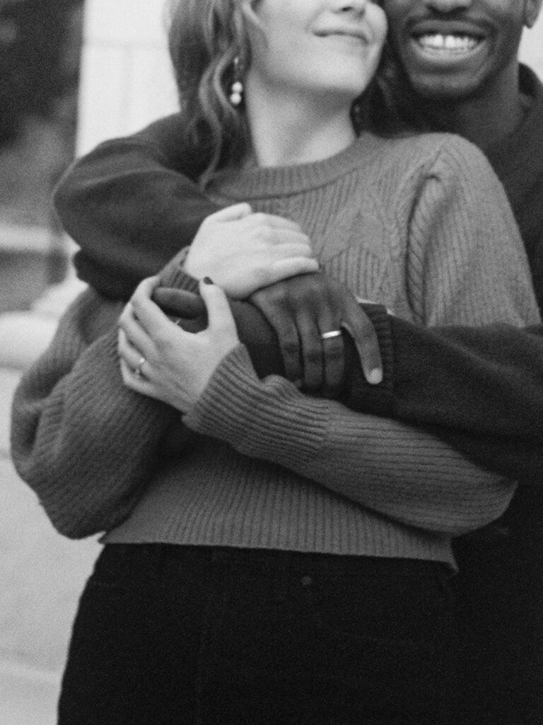 A close up photograph of a couple's hands, wrapped around each other. The guy is standing behind the girl, hugging her from behind. Her arms are wrapped around his.