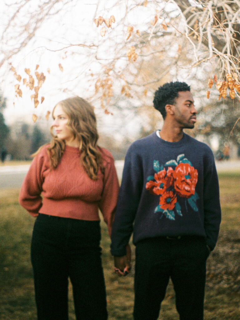 A couple stands in a park under a tree with brown leaves. They are holding hands, and looking in opposite directions.