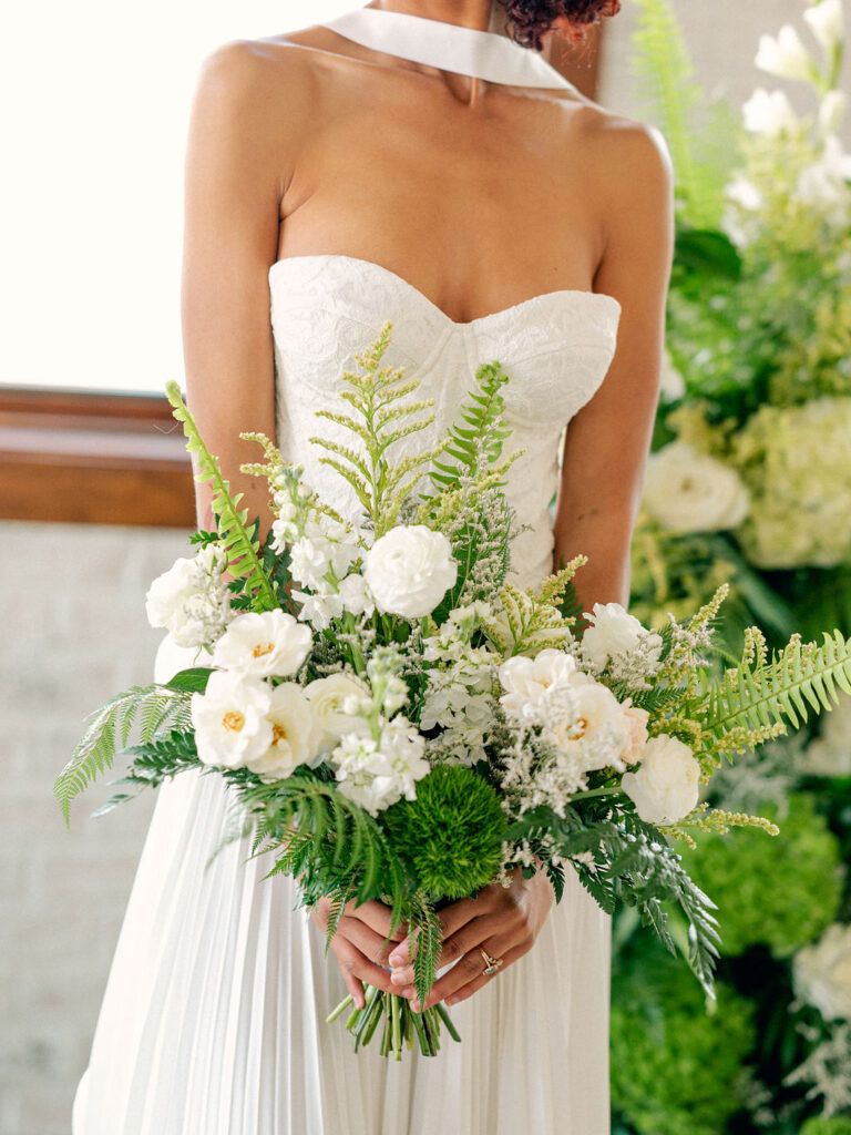 A bride holds in a modern drop-waist wedding gown with a neck scarf holds a bridal bouquet of greenery and white florals.