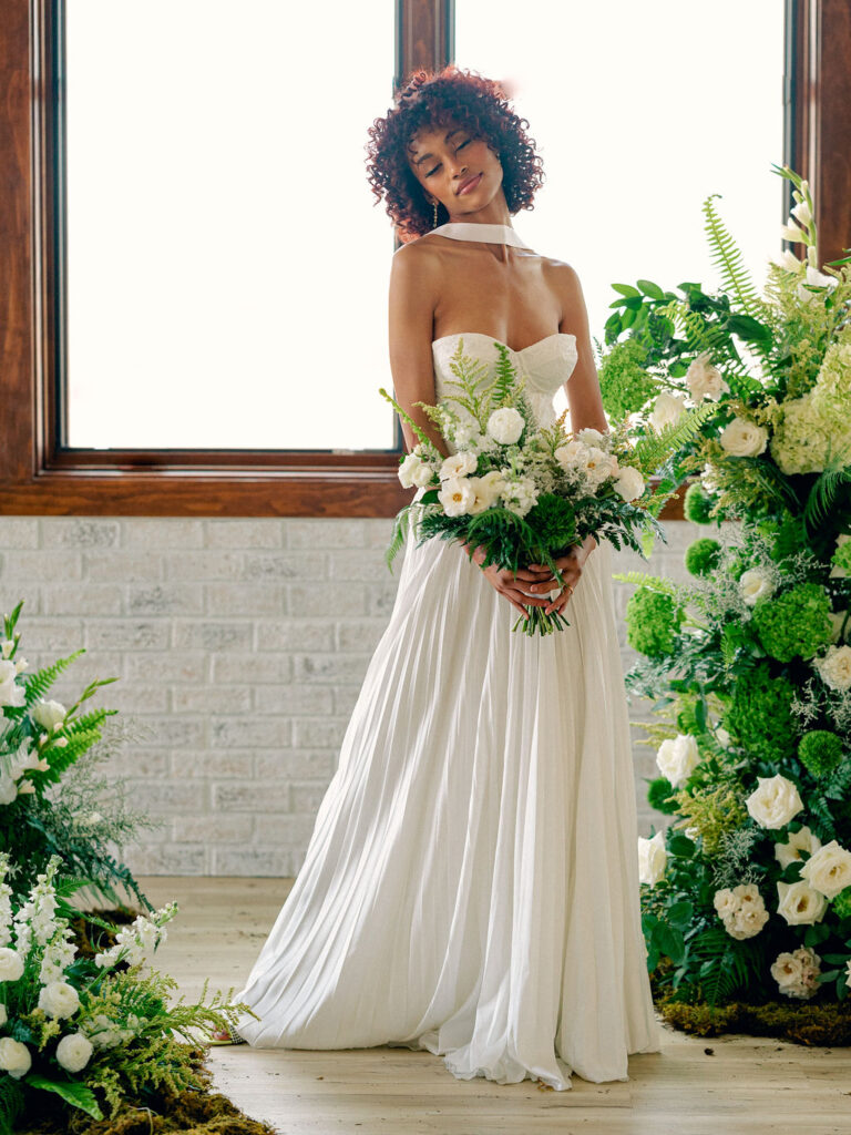 A bride in a modern drop waist gown with a neck scarf stands among a floral installation of greenery and white florals.