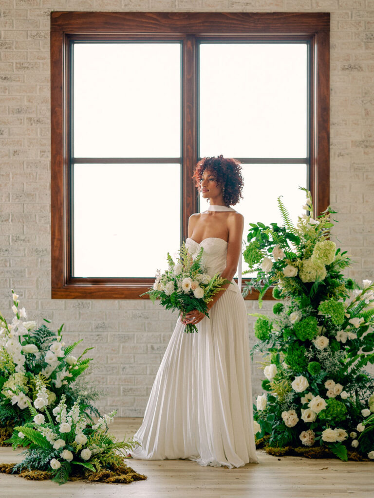 A bride in a drop waist wedding gown stands in front of a dramatic wedding ceremony floral installation of greenery and white florals.